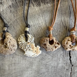 Könnte beinhalten: Vier einzigartige Steinanhänger an Lederbändern. Die Anhänger haben verschiedene Formen und Größen mit natürlichen Löchern und Texturen. Die Kordeln sind schwarz und braun, mit silberfarbenen Metallakzenten. Präsentiert auf einer verwitterten Holzoberfläche.