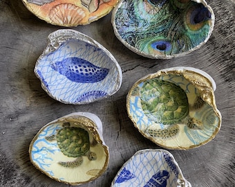 May include: Six decorative seashell dishes with silver and gold trim. The shells feature various designs, including a peacock feather pattern, a turtle, and other sea-themed illustrations. The shells are arranged on a rustic wooden surface.
