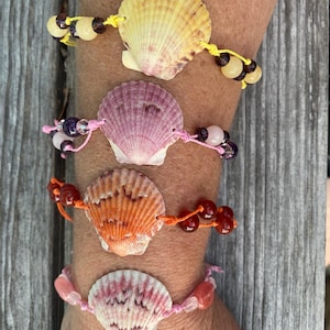 Puede incluir: Cuatro pulseras de conchas marinas con cuentas de diferentes colores. Las conchas son amarillas, rosas, naranjas y blancas con detalles rosas. Las pulseras están atadas con hilo amarillo, rosa y naranja. Las pulseras están en un brazo.