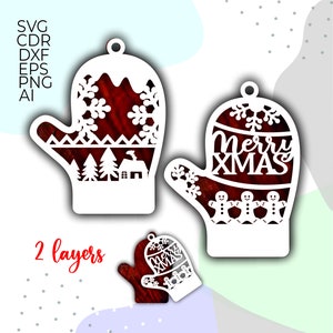 Op de afbeelding: Twee witte, uit papier gesneden kerstmanchet-versieringen. Eén manchet heeft een rode achtergrond met sneeuwvlokken en een klein huis met bomen. De andere manchet heeft een rode achtergrond met de tekst "Merry Xmas" en peperkoekmannetjes.