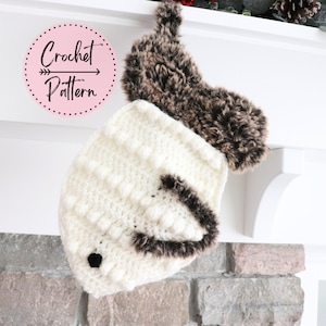 May include: White crocheted stocking with a brown faux fur trim and a black nose. The stocking is hanging on a white mantel above a brick fireplace. The text "Crochet Pattern" is written in a pink circle.