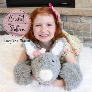 May include: A grey crocheted bunny lovey with pink flower accents. The lovey is medium sized and is being held by a young girl with red hair. The image includes the text "Crochet Pattern" and "Lovey Size: Medium".