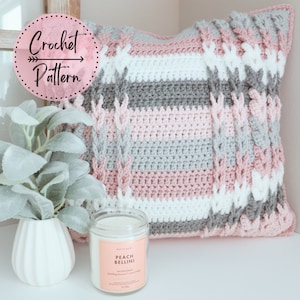 May include: A crocheted pillow with a cable knit pattern in shades of pink, gray, and white. The pillow is sitting on a white surface with a candle in front of it. The candle is labeled "Peach Bellini" and has a pink label.