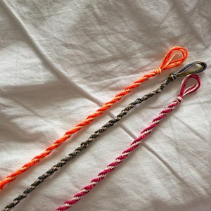 May include: Three twisted cord bracelets, each featuring a loop at one end. The first is bright orange and yellow, the second is a mix of dark blue, green, and brown, and the third is a combination of vibrant pink and white thread strands.