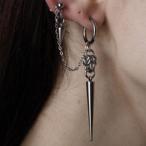 Può includere: Set di orecchini doppi color argento. Un orecchino presenta una catena che collega un piccolo cono a un perno. L'altro orecchino ha un cerchio con una catena e un lungo pendente a cono appuntito.