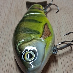 May include: A detailed fishing lure with a fish-like design. The lure features a green and yellow body with brown accents, a large eye, and multiple hooks. The lure is designed to attract fish.