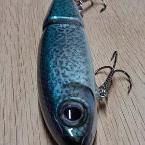 May include: A detailed close-up of a fishing lure shaped like a fish. The lure is primarily blue and silver with a textured pattern, featuring a realistic eye and two treble hooks. The lure is designed to mimic a baitfish.