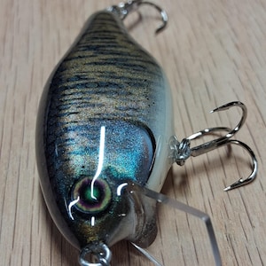 Puede incluir: Un señuelo de pesca con acabado metálico, que presenta un diseño detallado con tonos de azul, plata y oro. Tiene un labio de plástico transparente y dos anzuelos triples. El señuelo está diseñado para atraer peces.