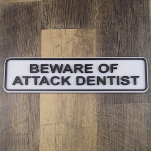 Puede incluir: Un letrero rectangular blanco con texto negro que dice "BEWARE OF ATTACK DENTIST". El letrero tiene un borde negro y está sobre un fondo de madera.