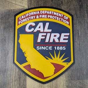 Könnte beinhalten: Ein schildförmiges Emblem für das kalifornische Ministerium für Forstwirtschaft und Brandschutz. Das Emblem hat eine gelbe Umrandung, einen blauen Hintergrund und die Worte "CAL FIRE" in Weiß. Die Form des Bundesstaates Kalifornien ist gelb, und das Emblem lautet "SEIT 1885".