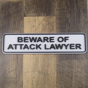 May include: A rectangular white sign with black text that reads "BEWARE OF ATTACK LAWYER." The sign has a black border and is set against a wooden background. The text is in a bold, sans-serif font.