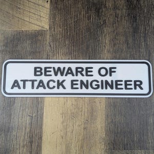 Puede incluir: Un letrero rectangular blanco con texto negro que dice "Beware of Attack Engineer".