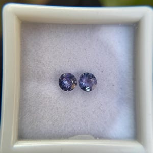 May include: Two round, purple gemstones in a white box. The gemstones are approximately 5mm in diameter.