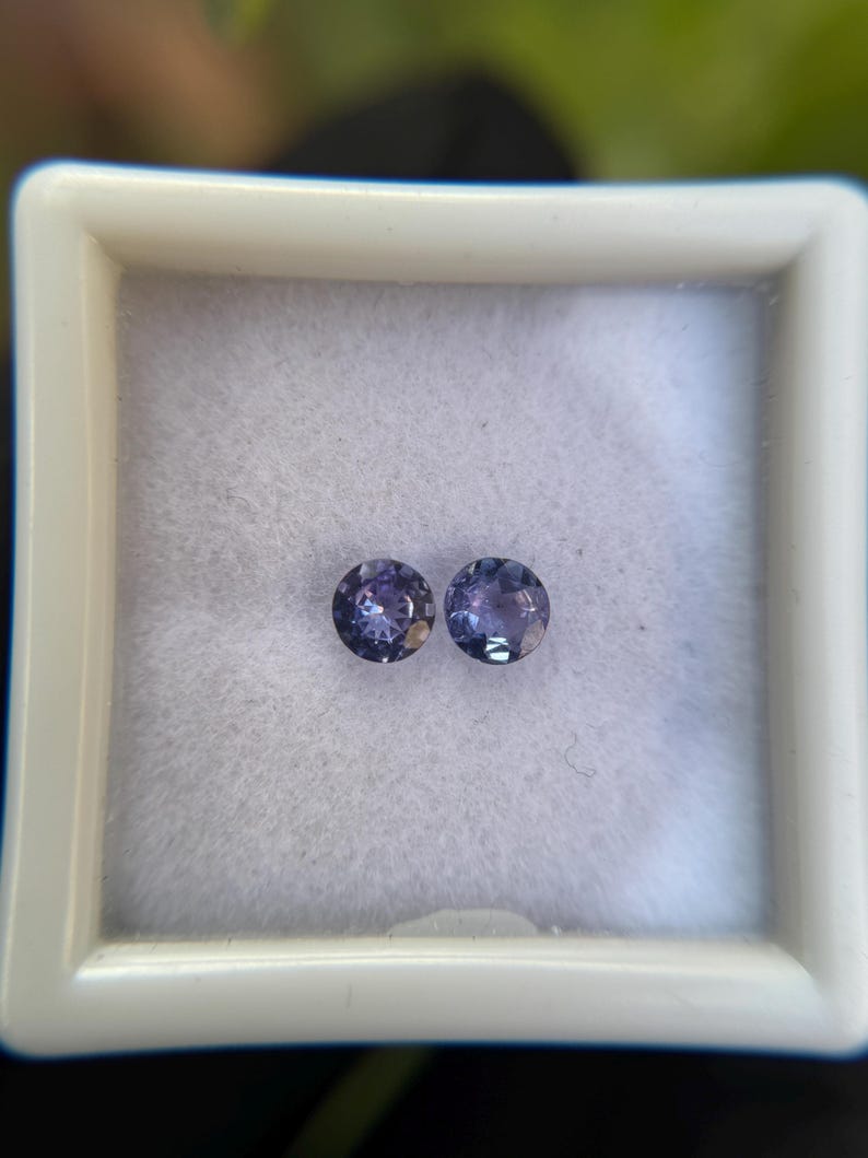 May include: Two round, purple gemstones in a white box. The gemstones are approximately 5mm in diameter.