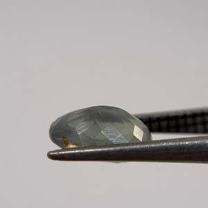 May include: A faceted, oval-shaped gemstone in shades of gray and green is held by a pair of metal tweezers. The gem has multiple facets that reflect light, creating a sparkling effect. The background is a soft, neutral color.
