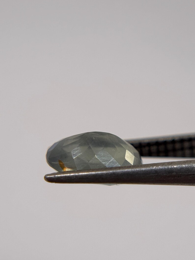 May include: A faceted, oval-shaped gemstone in shades of gray and green is held by a pair of metal tweezers. The gem has multiple facets that reflect light, creating a sparkling effect. The background is a soft, neutral color.