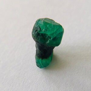 May include: A dark green emerald crystal with a rough, faceted surface. The crystal is partially covered in a black matrix.