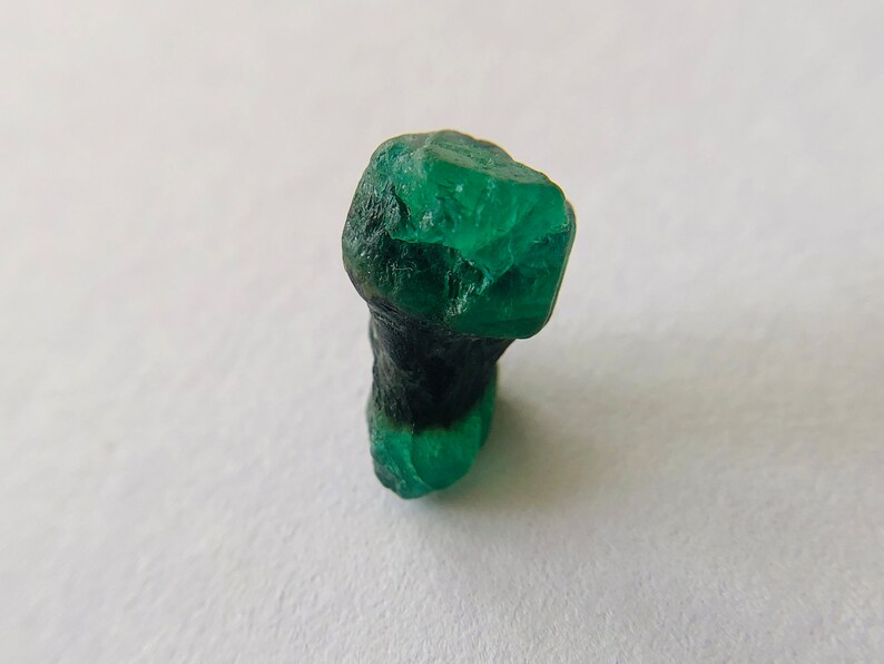 May include: A dark green emerald crystal with a rough, faceted surface. The crystal is partially covered in a black matrix.