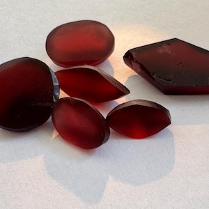 May include: Several translucent, deep red gemstones in different shapes. The stones include oval, rounded, and geometric cuts, displaying a rich, dark colour. The gems are arranged on a white surface, with light reflecting off their polished surfaces.