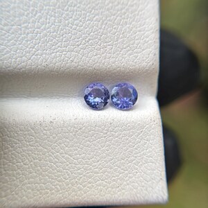 May include: Two round, purple gemstones on a white, textured surface.