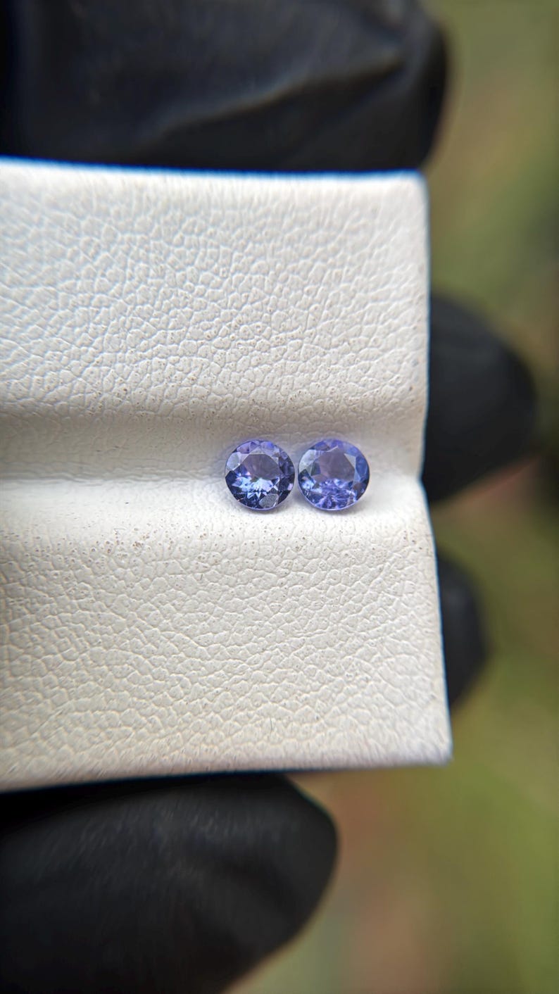 May include: Two round, purple gemstones on a white, textured surface.