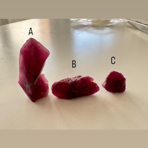 Może przedstawiać: Trzy surowe, nieoszlifowane kamienie szlachetne rubinu o różnych rozmiarach. Rubiny mają głęboki, bogaty czerwony kolor, z szorstką, teksturowaną powierzchnią. Kamienie szlachetne są prezentowane na jasnej powierzchni.