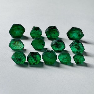 Könnte beinhalten: Eine Sammlung von 13 smaragdgrünen Edelsteinen, jeder mit einer sechseckigen Form und einer leicht unterschiedlichen Größe und einem leicht unterschiedlichen Schliff. Die Edelsteine sind in einer lockeren Gruppe auf einer weißen Oberfläche angeordnet.