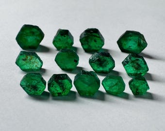 May include: A collection of 13 emerald green gemstones, each with a hexagonal shape and a slightly different size and cut. The gemstones are arranged in a loose cluster on a white surface.