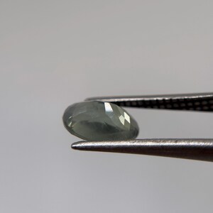 May include: A faceted, oval-shaped gemstone in shades of gray and green is held by a pair of tweezers. The gem has multiple facets that reflect light, creating a sparkling effect. The background is a plain, light gray.