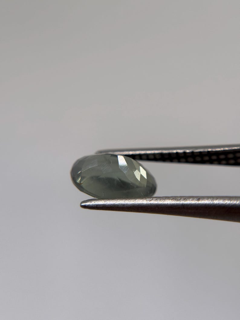 May include: A faceted, oval-shaped gemstone in shades of gray and green is held by a pair of tweezers. The gem has multiple facets that reflect light, creating a sparkling effect. The background is a plain, light gray.