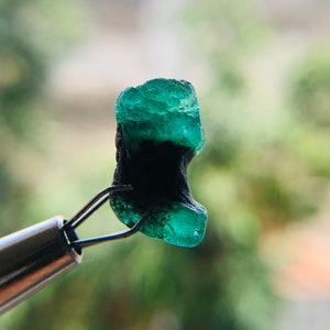May include: A dark green emerald gemstone with a black inclusion, held by a pair of tweezers.