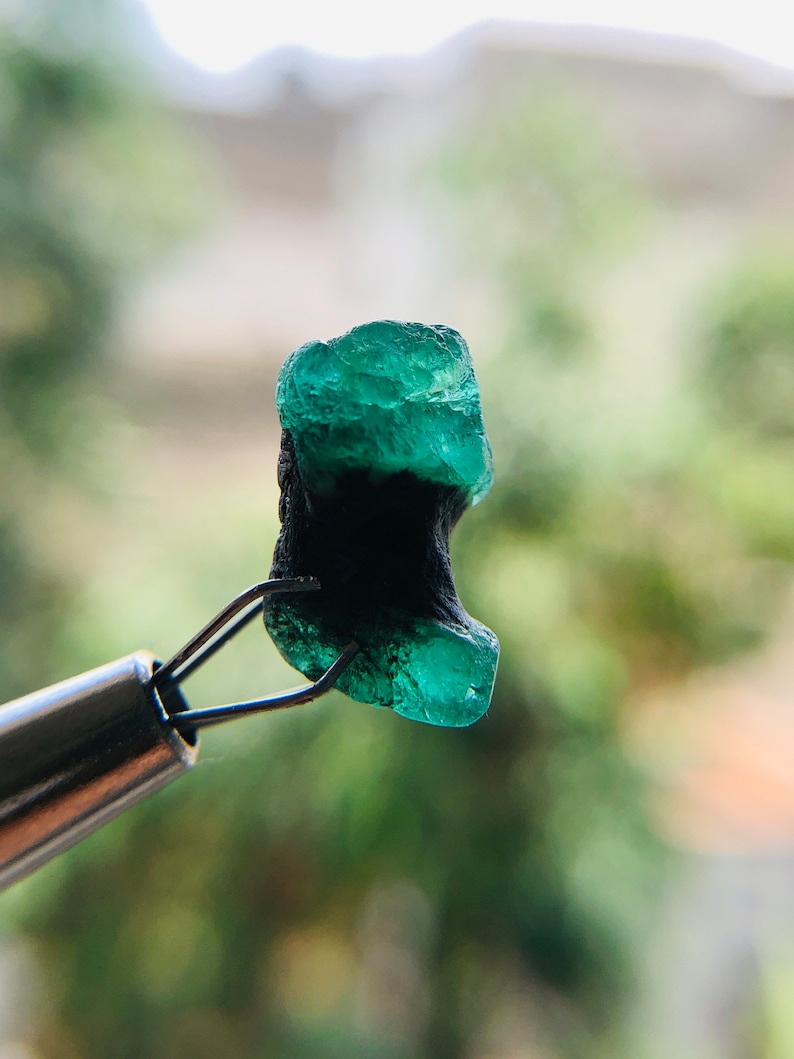 May include: A dark green emerald gemstone with a black inclusion, held by a pair of tweezers.