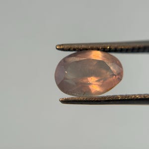 May include: A faceted, oval-shaped gemstone held by tweezers. The gem has a peach and lavender color with internal light reflections. The tweezers are a dark metal, and the background is a soft gray.