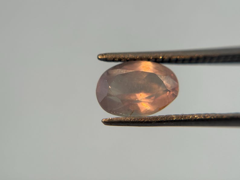 May include: A faceted, oval-shaped gemstone held by tweezers. The gem has a peach and lavender color with internal light reflections. The tweezers are a dark metal, and the background is a soft gray.