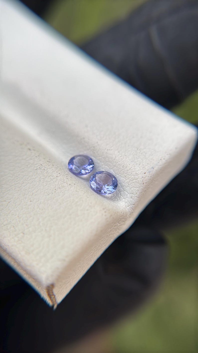 May include: Two small, round, purple gemstones on a white surface.