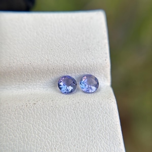 May include: Two round, purple gemstones on a white, textured surface.
