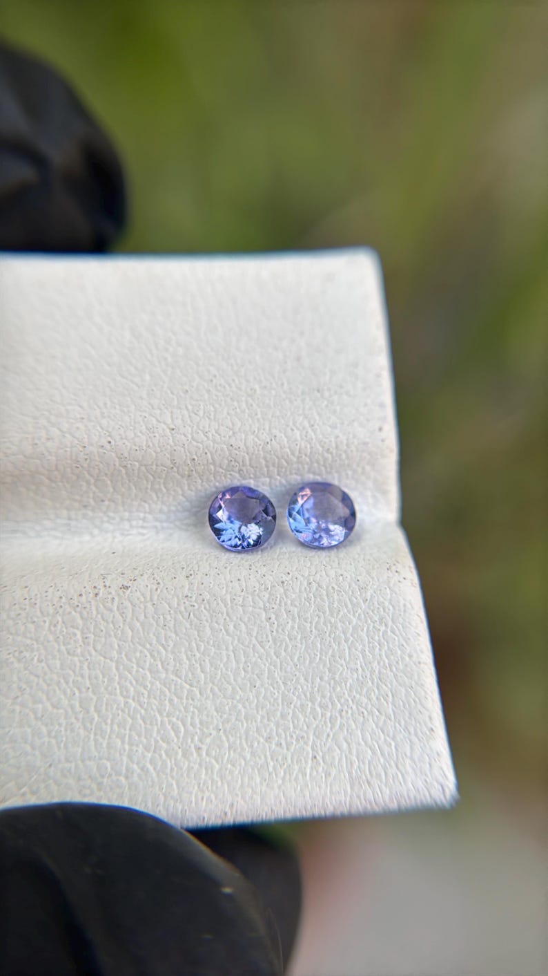 May include: Two round, purple gemstones on a white, textured surface.