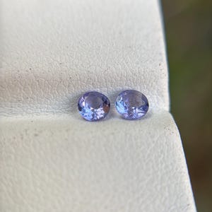 May include: Two round, purple gemstones on a white surface.