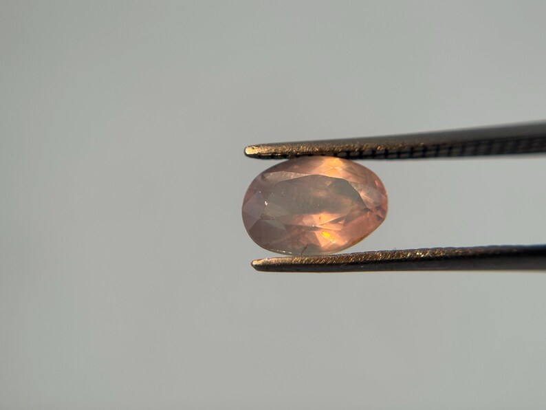 May include: An oval-shaped, peach-colored gemstone is held by a pair of tweezers. The gem has a translucent quality, with hints of orange and yellow light reflecting within its facets. The tweezers are dark, contrasting with the gem.