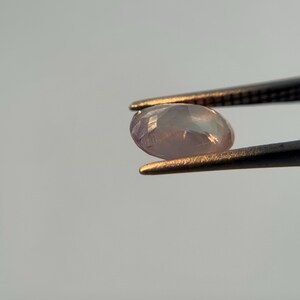 May include: A faceted, oval-shaped gemstone, held by tweezers. The gem has a pale pink hue with iridescent flashes. The tweezers are black with gold-colored tips, set against a light gray background.