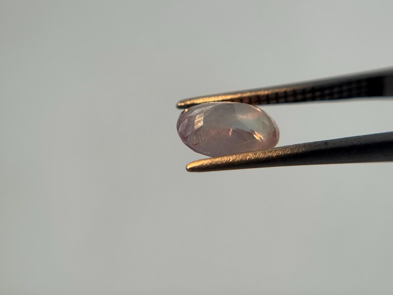 May include: A faceted, oval-shaped gemstone, held by tweezers. The gem has a pale pink hue with iridescent flashes. The tweezers are black with gold-colored tips, set against a light gray background.