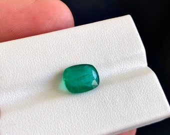 May include: A vibrant green emerald gemstone, cut into a cushion shape, displayed on a white background.