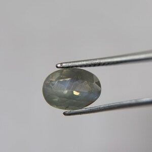 May include: A faceted, oval-shaped gemstone held by silver tweezers. The gem has a pale, translucent color with subtle internal reflections. The tweezers are positioned to display the gem against a neutral background, highlighting its clarity and cut.