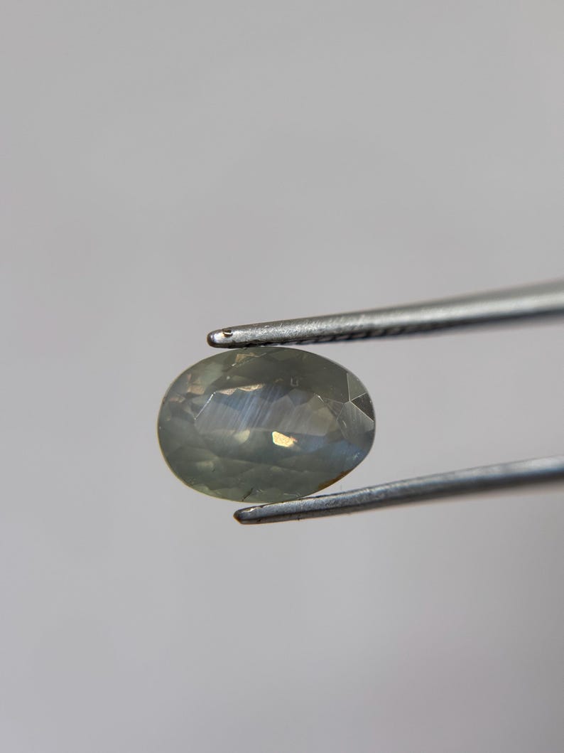 May include: A faceted, oval-shaped gemstone held by silver tweezers. The gem has a pale, translucent color with subtle internal reflections. The tweezers are positioned to display the gem against a neutral background, highlighting its clarity and cut.