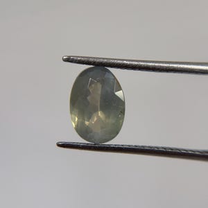 May include: An oval-shaped, pale green gemstone is held by a pair of metal tweezers against a neutral background. The gemstone has multiple facets, reflecting light. The tweezers are dark gray.