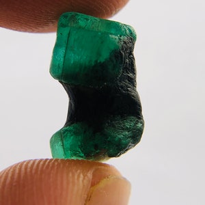 May include: A dark green emerald gemstone with a rough, natural shape. The emerald is partially covered in a black, opaque material.