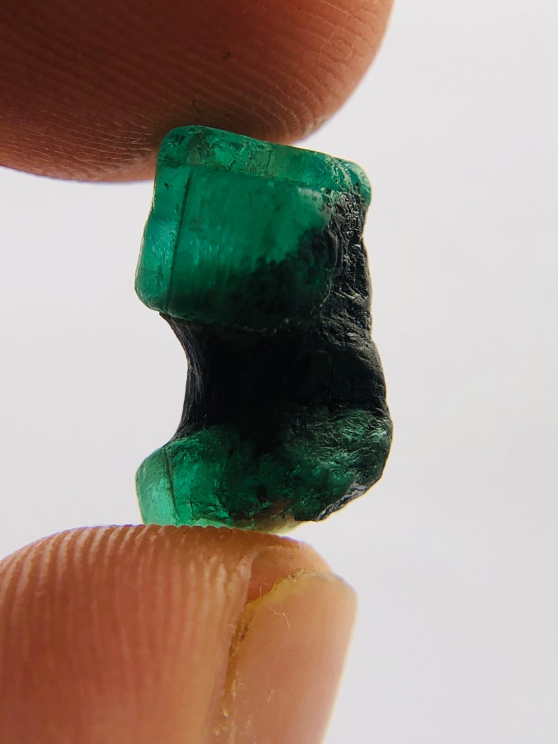 May include: A dark green emerald gemstone with a rough, natural shape. The emerald is partially covered in a black, opaque material.