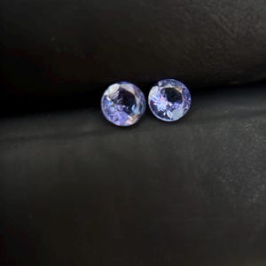 May include: Two round, purple gemstones with a faceted cut. The gemstones are set against a dark gray background.