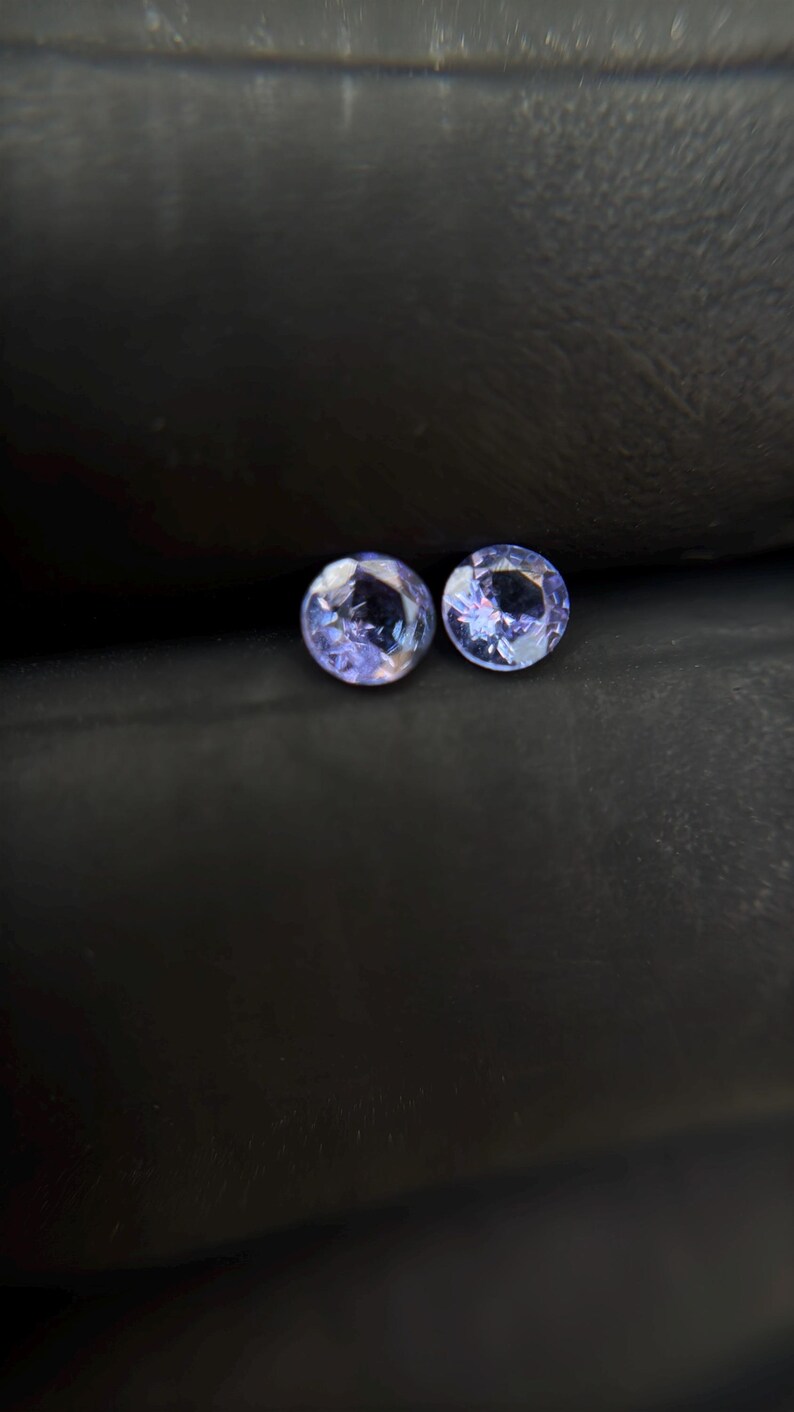 May include: Two round, purple gemstones with a faceted cut. The gemstones are set against a dark gray background.