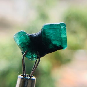 May include: A dark green emerald gemstone with a rough, faceted surface. The emerald is held in place by a pair of metal tweezers.